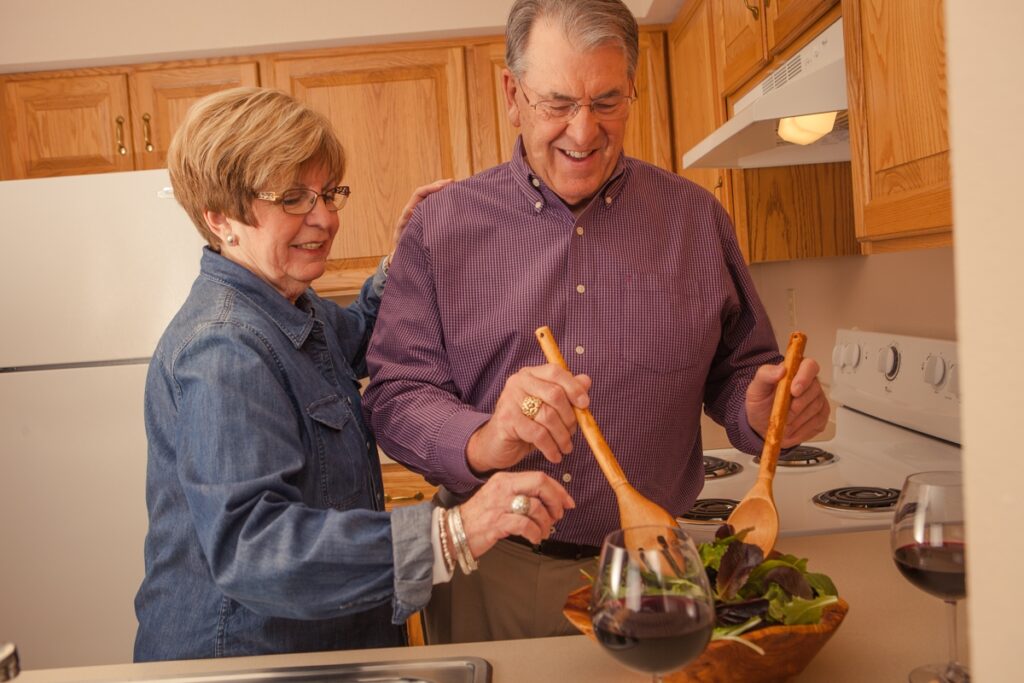 Older couple smiling mixing a salad and two glasses of wine sitting on the counter