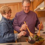 Older couple smiling mixing a salad and two glasses of wine sitting on the counter