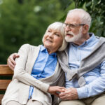 smiling senior couple embracing while sitting on wooden bench in park